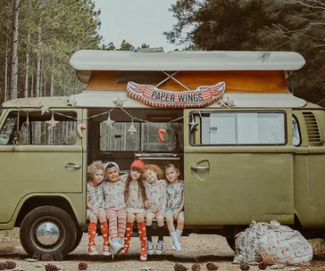 Kids in festive outfits sitting in a vintage van with "Paper Wings" sign, surrounded by trees and decorations.