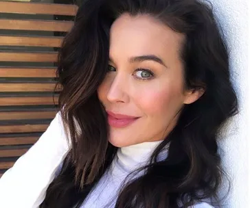 Portrait of a woman with long dark hair, smiling at the camera, wearing a white top.