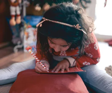 A girl in a festive sweater writes a letter with a candy cane-striped pencil, sitting cross-legged on a cushion indoors.
