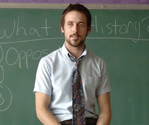 Man in a shirt and tie sits on desk in front of a chalkboard with writing.