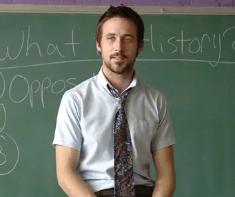 Man in a shirt and tie sits on desk in front of a chalkboard with writing.
