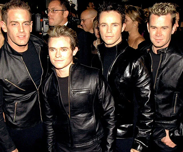 Four men in black leather jackets pose together at an event, surrounded by other people.