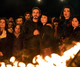 A group of people stand together at night, gathered around a warm, glowing fire, looking contemplative.