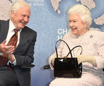 Two elderly individuals, dressed formally, sit and share a light-hearted moment with smiles against a world map backdrop.