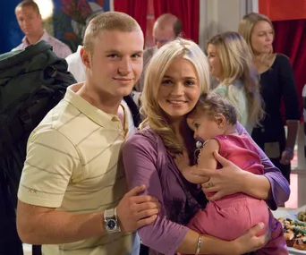 A young couple holds a baby while smiling at a social gathering, with people chatting in the background.