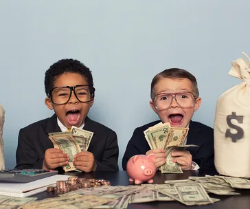 Two kids in suits excitedly holding cash, with coins, a piggy bank, and a money bag on the table.