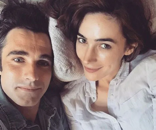 A couple lying close together, smiling at the camera with heads resting on a pillow in a cozy setting.