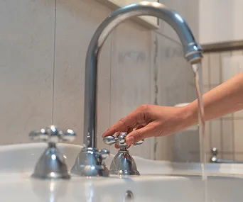 Hand turning off a bathroom tap with water flowing, illustrating water conservation measures.