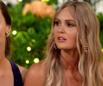 Two women seated; one with long blonde hair is looking to the side, with a blurred garden background.