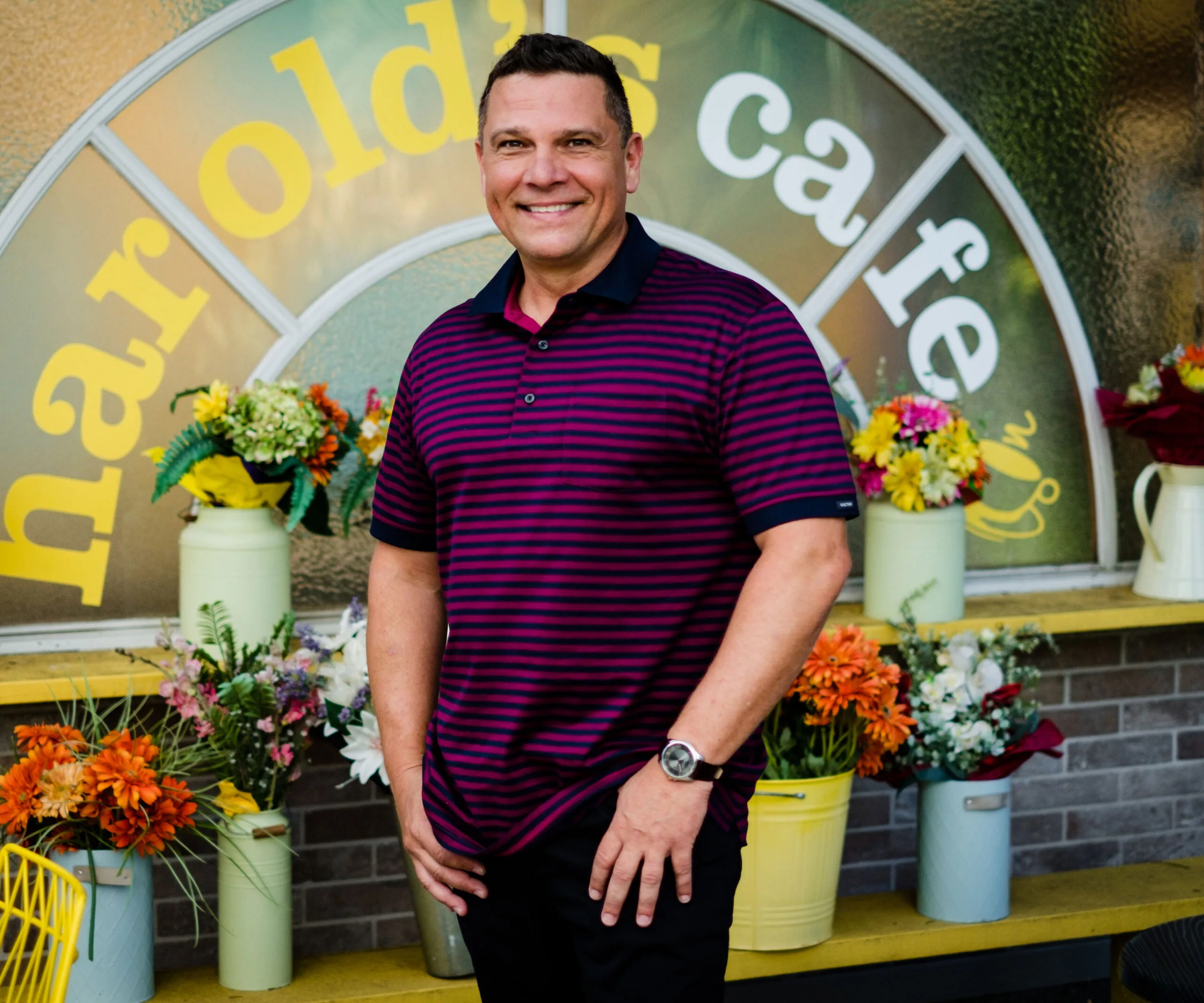 Man smiling in front of Harold's Café window, surrounded by colorful flowers.