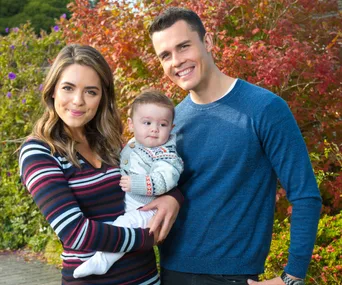 A smiling couple outdoors with a baby in a cozy outfit, set against colorful foliage.