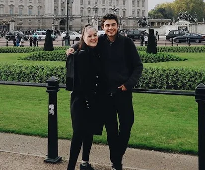 A smiling couple in black coats pose arm-in-arm in front of a palace with cars and gardens visible in the background.