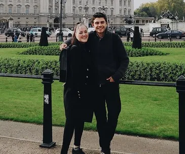 A smiling couple in black coats pose arm-in-arm in front of a palace with cars and gardens visible in the background.