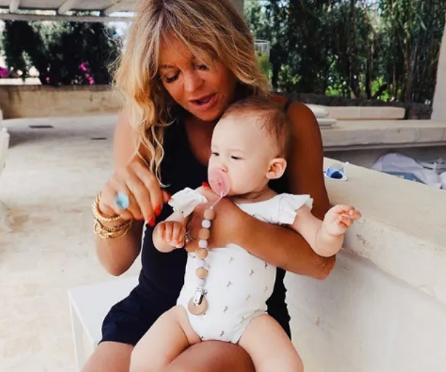 Woman with long hair holding a baby on her lap, baby with a pacifier, sitting outdoors.