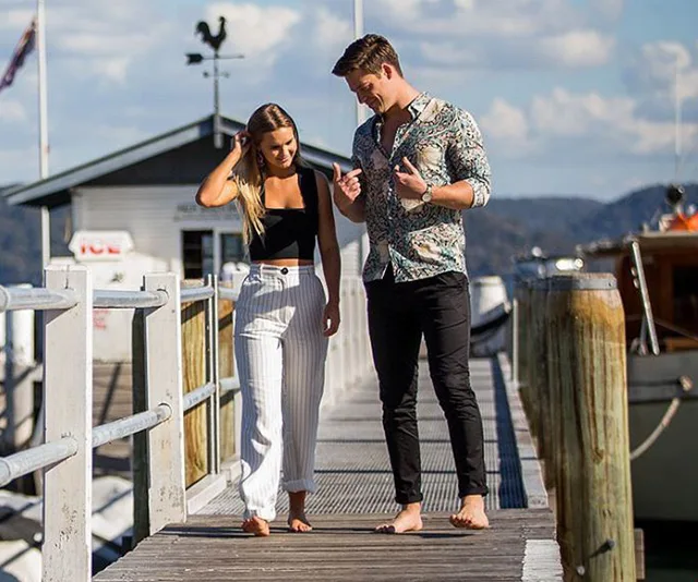 A couple walking barefoot on a wooden pier with a scenic view in the background.