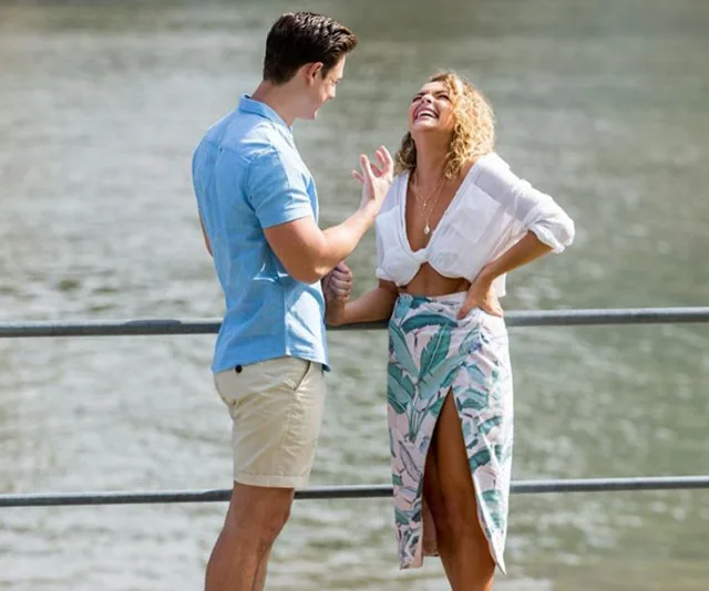 Man and woman laughing by the water, woman in printed skirt and white blouse, man in blue shirt and shorts.