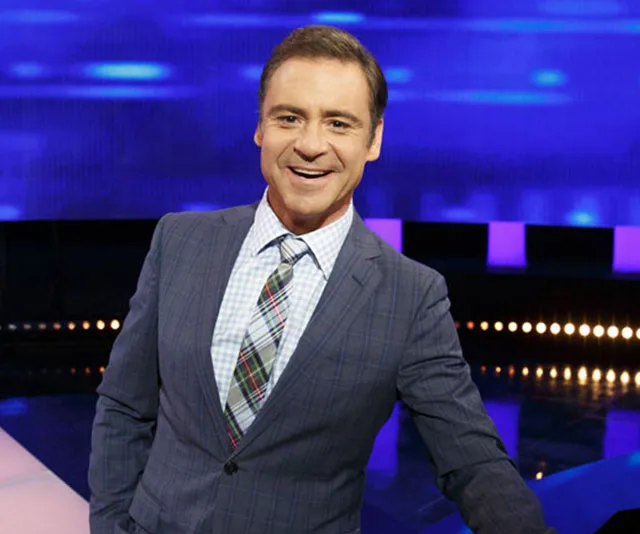 Presenter smiling on a TV game show set with blue and purple lighting.