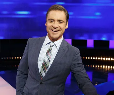 Presenter smiling on a TV game show set with blue and purple lighting.