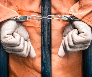 Inmate in orange jumpsuit with hands cuffed behind bars.