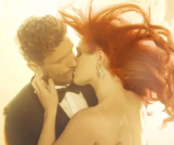 A couple passionately kissing, with the woman's striking red hair flowing dramatically, set against a luminous background.