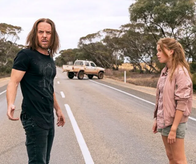 Man and woman stand on a rural road with a pickup truck in the background, surrounded by trees.