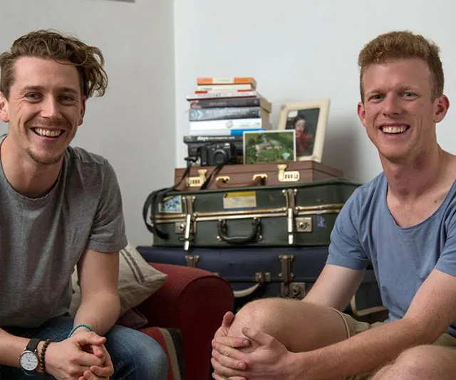 Two smiling men are seated in a cozy room with stacked suitcases and books in the background.
