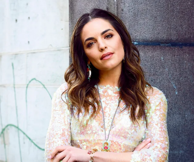 Woman in a floral dress leans confidently against a wall, with long wavy hair and accessorized with earrings and a necklace.