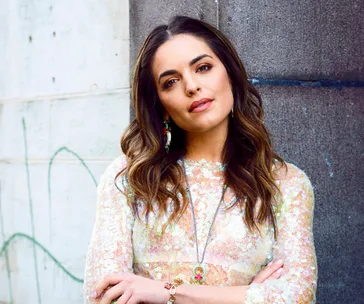 Woman in a floral dress leans confidently against a wall, with long wavy hair and accessorized with earrings and a necklace.