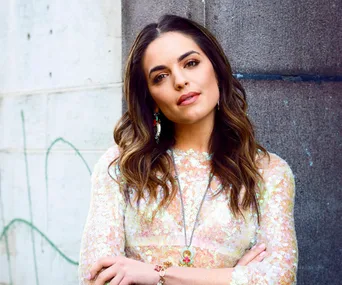 Woman in a floral dress leans confidently against a wall, with long wavy hair and accessorized with earrings and a necklace.
