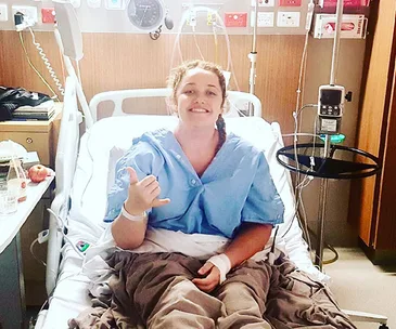 Person in a hospital bed smiling, wearing a hospital gown, surrounded by medical equipment and giving a thumbs-up.
