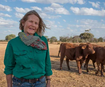 A farmer in a green shirt stands in a dry paddock with cattle, under a partly cloudy sky.