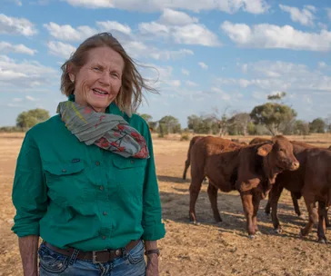 A farmer in a green shirt stands in a dry paddock with cattle, under a partly cloudy sky.