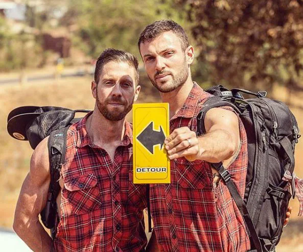 Two men in plaid shirts hold a "Detour" sign on a hiking trail, wearing backpacks under a sunny sky.