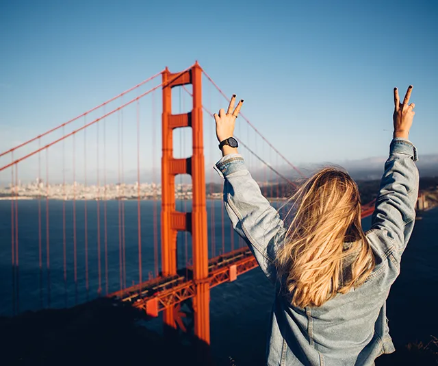 Blonde woman in denim jacket raising peace signs at Golden Gate Bridge in San Francisco.