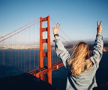 Blonde woman in denim jacket raising peace signs at Golden Gate Bridge in San Francisco.