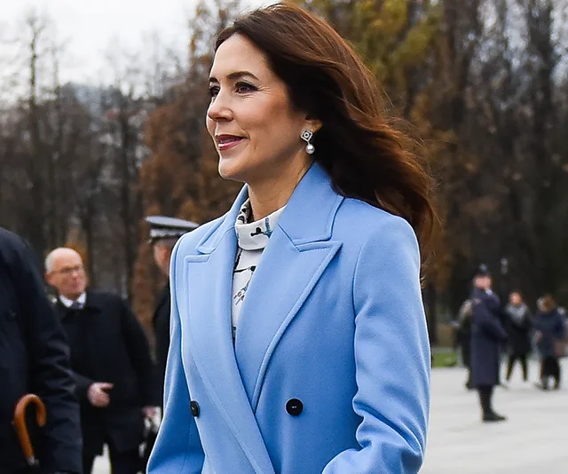 Princess Mary of Denmark in a light blue coat, smiling outdoors, with blurred people in the background.
