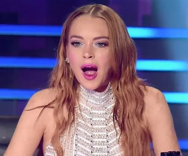 Lindsay Lohan appears surprised with mouth open, wearing a detailed white dress against a blue lit background.