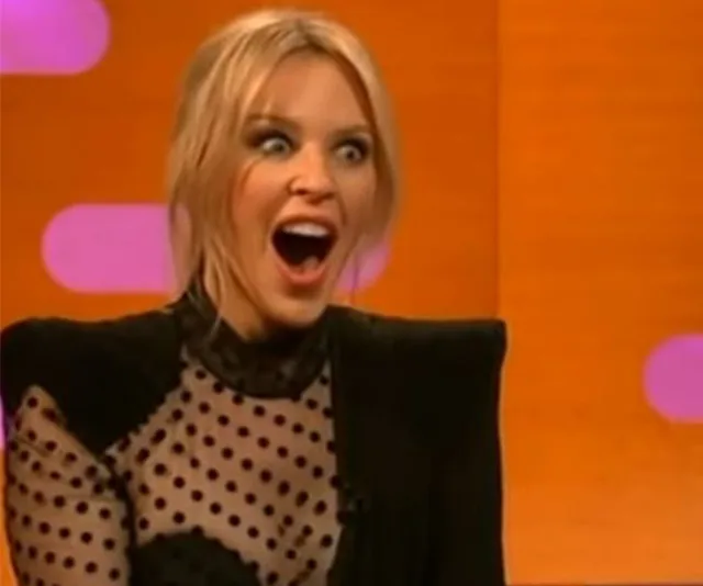 Woman with surprised expression on a TV talk show set, wearing a black polka dot top.