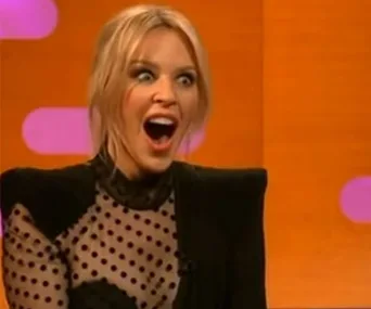 Woman with surprised expression on a TV talk show set, wearing a black polka dot top.