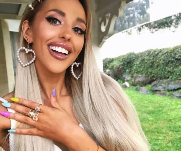 A woman with long blonde hair, wearing pearl heart earrings and colorful nails, smiles outdoors.