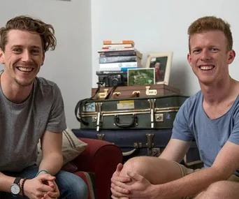 Two men smiling, sitting in a living room with books, a camera, and luggage stacked in the background.