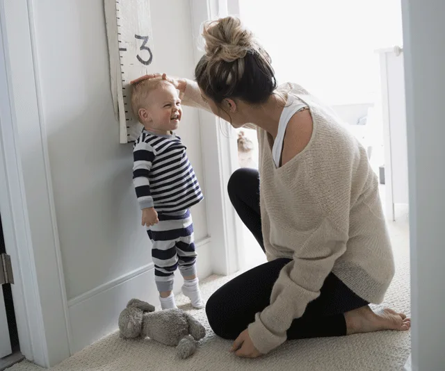 Mother measuring smiling toddler's height with a growth chart, stuffed toy on floor.