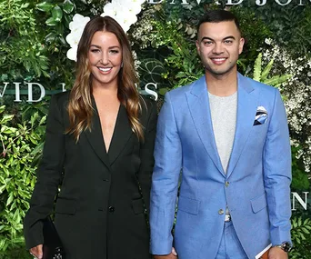 Two people smiling in formal attire; one wearing a dark blazer, the other in a blue suit. Greenery backdrop.