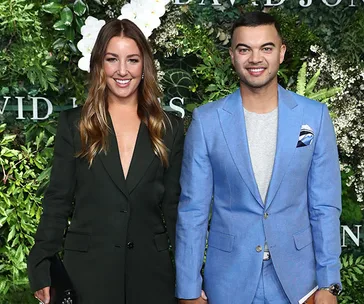 Two people smiling in formal attire; one wearing a dark blazer, the other in a blue suit. Greenery backdrop.