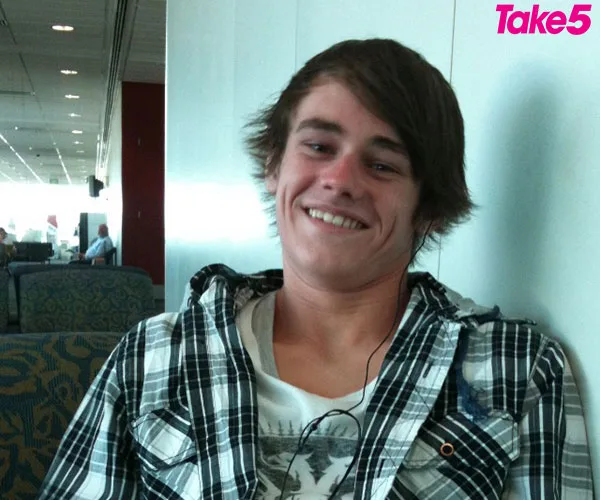 Young man smiling in a plaid shirt, seated in an airport lounge, with earphones.