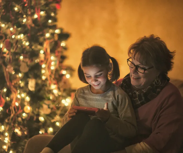 Grandparent and grandchild enjoying a tablet together by a decorated Christmas tree.