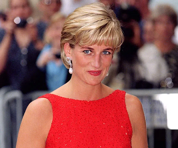 A woman in a red dress with short blonde hair and pearl earrings, smiling at a public event.