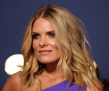 Blonde woman in a purple dress with wavy hair against a dark background.