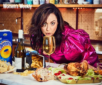Woman in pink blouse drinking from a glass at a table with snacks, wine, and roasted chicken; magazine logo in corner.