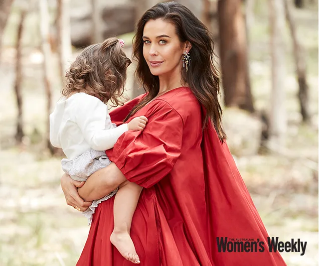 Woman in a flowing red dress holds a child in a forest setting, both looking serene, featured in Women's Weekly magazine.